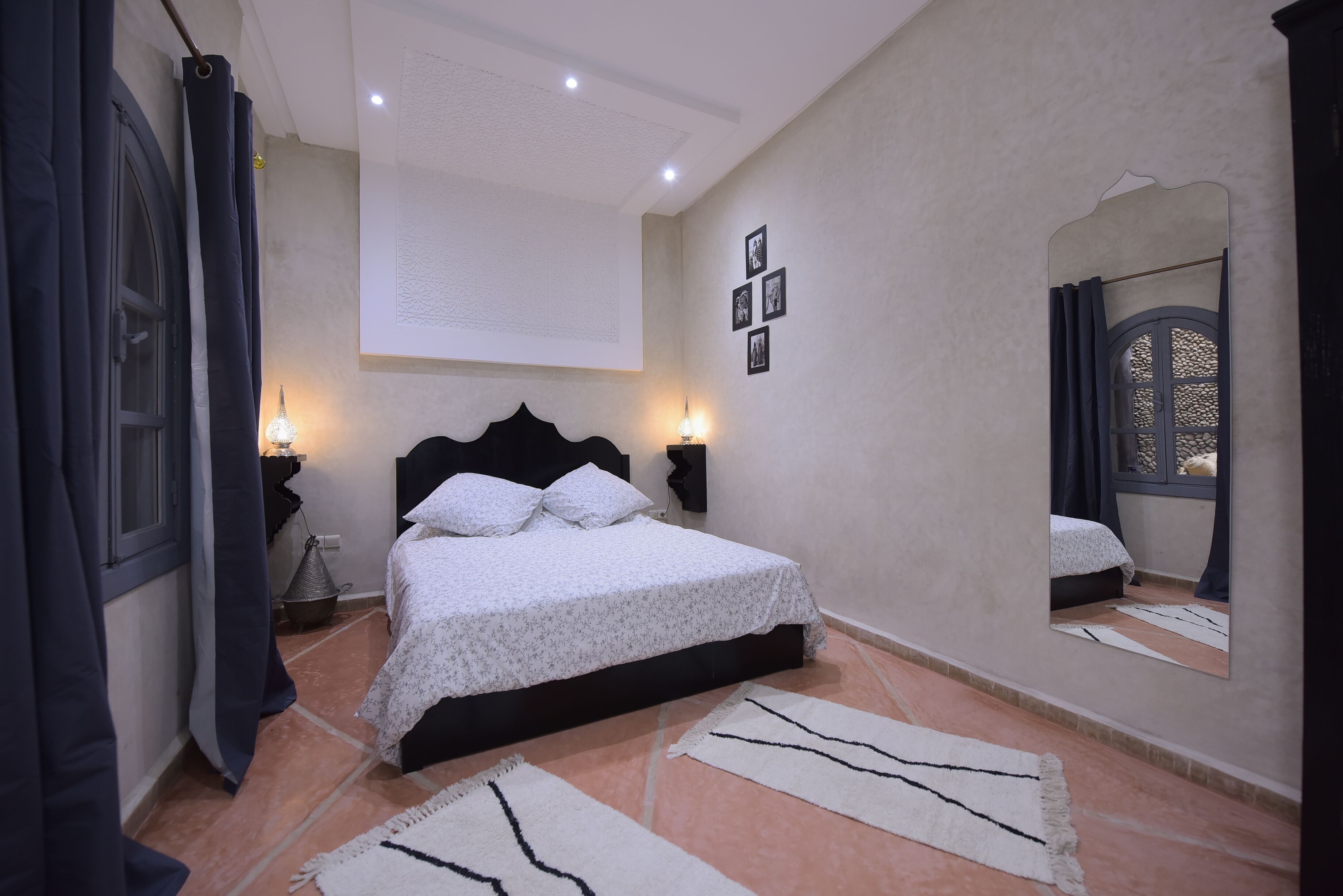 Un dormitorio con cama, lámparas de noche, espejo en la pared y ventana con cortinas.