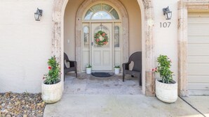 Une entrée d’immeuble avec une porte beige, une guirlande, deux plantes en pot et le numéro « 107 » sur le mur.