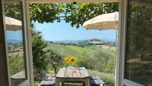 Eine Terrasse mit Holztisch und -stühlen, einer Vase mit Sonnenblumen und Blick auf sanfte Hügel und Berge.