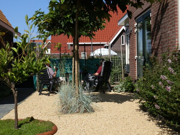 A courtyard with a gravel path, two chairs, a small tree, and a brick building with a red roof in the background.