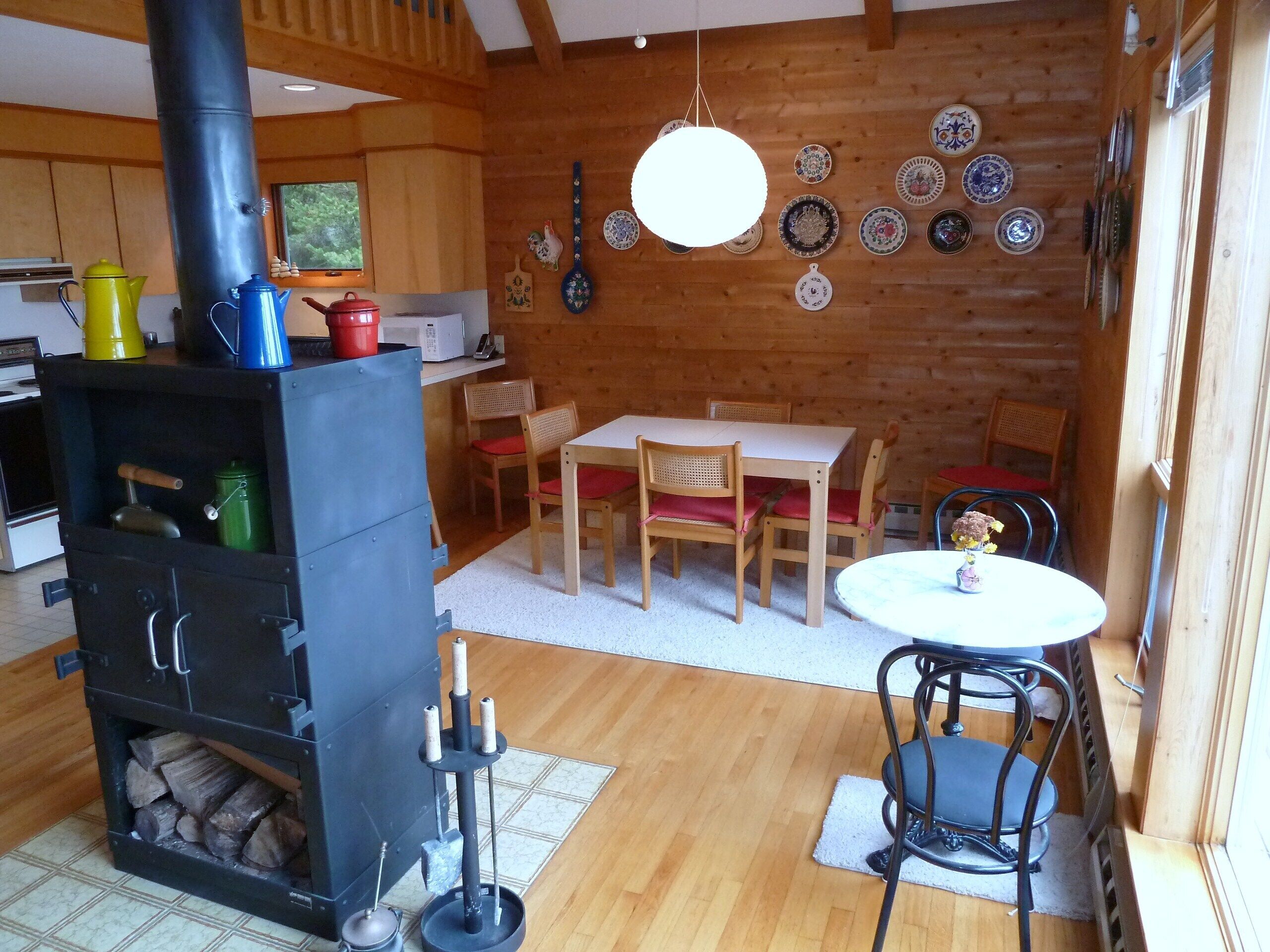 A cozy cabin interior with a wood stove, dining table, chairs, and a variety of decorative plates on the wall.