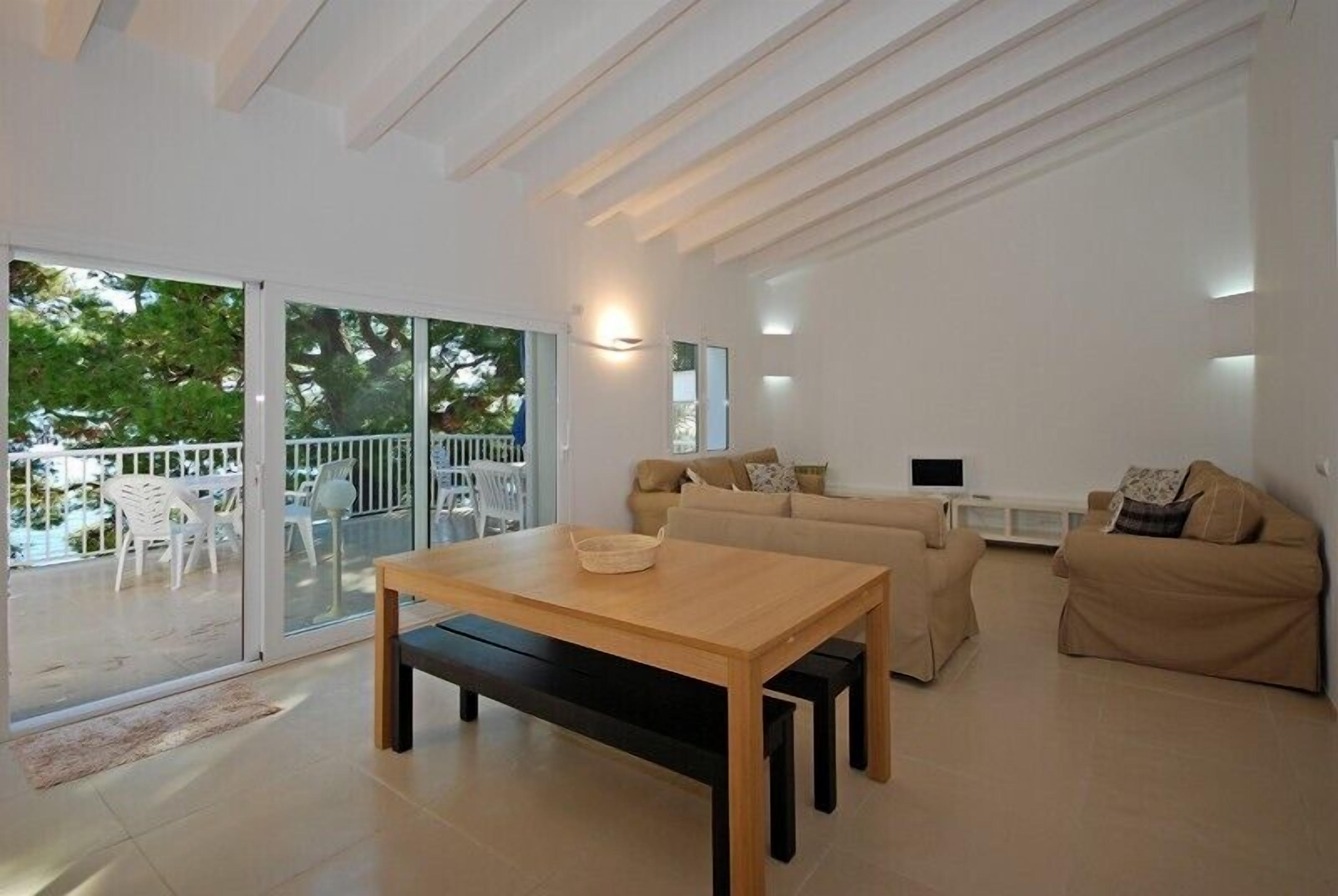 A modern living room with a wooden table, beige sofas, and a glass door leading to an outdoor area with white chairs.