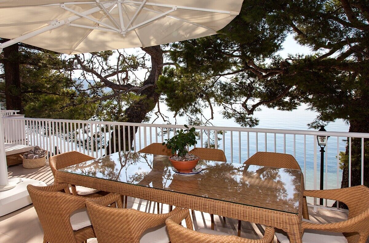 A glass-top table with wicker chairs and a potted plant, set against a backdrop of trees and a body of water.