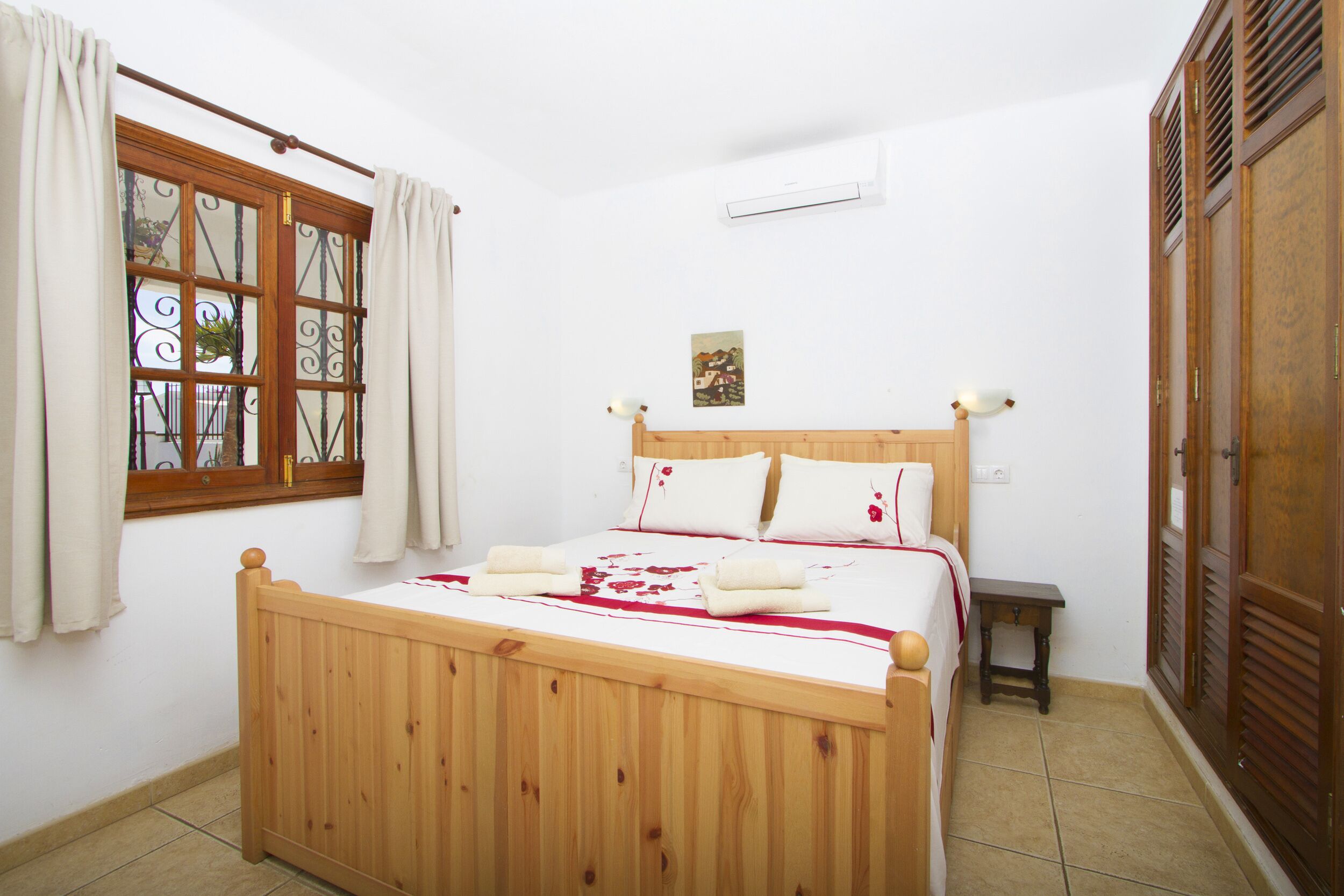 Ein Bett mit Holzbettgestell und weißer Bettwäsche, ein kleiner Nachttisch, ein Fenster mit Vorhängen und eine Wand-Klimaanlage.