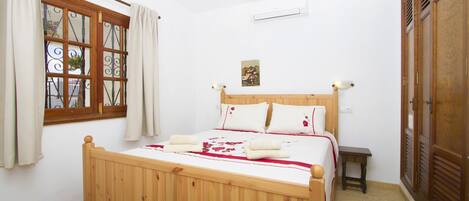 Ein Bett mit Holzbettgestell und weißer Bettwäsche, ein kleiner Nachttisch, ein Fenster mit Vorhängen und eine Wand-Klimaanlage.