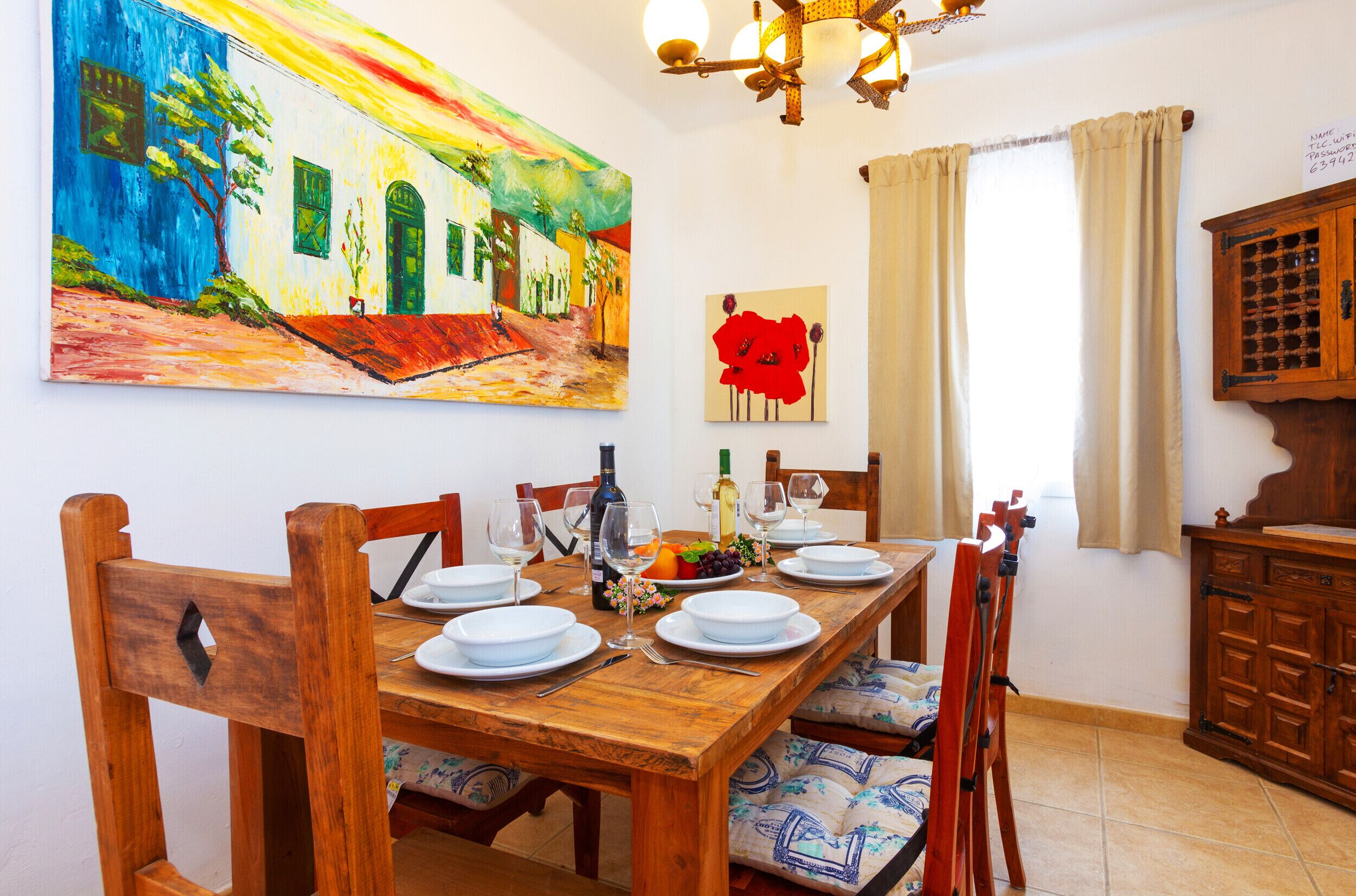 A dining area with a wooden table set for four, surrounded by chairs. A colorful painting and a red abstract artwork are on the wall.