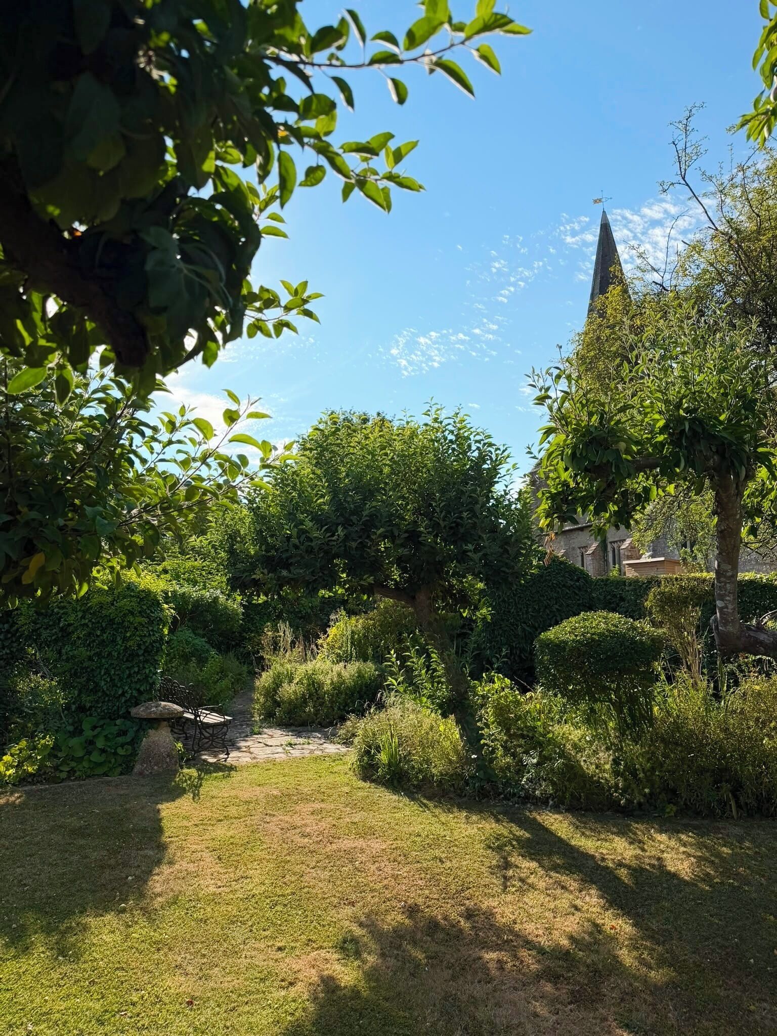 A garden with a stone pathway leading to a bench, surrounded by lush greenery and a church spire in the background.