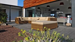 Un espace de vie extérieur moderne comprenant un canapé d’angle, une terrasse en bois et une porte vitrée donnant accès à un espace intérieur.