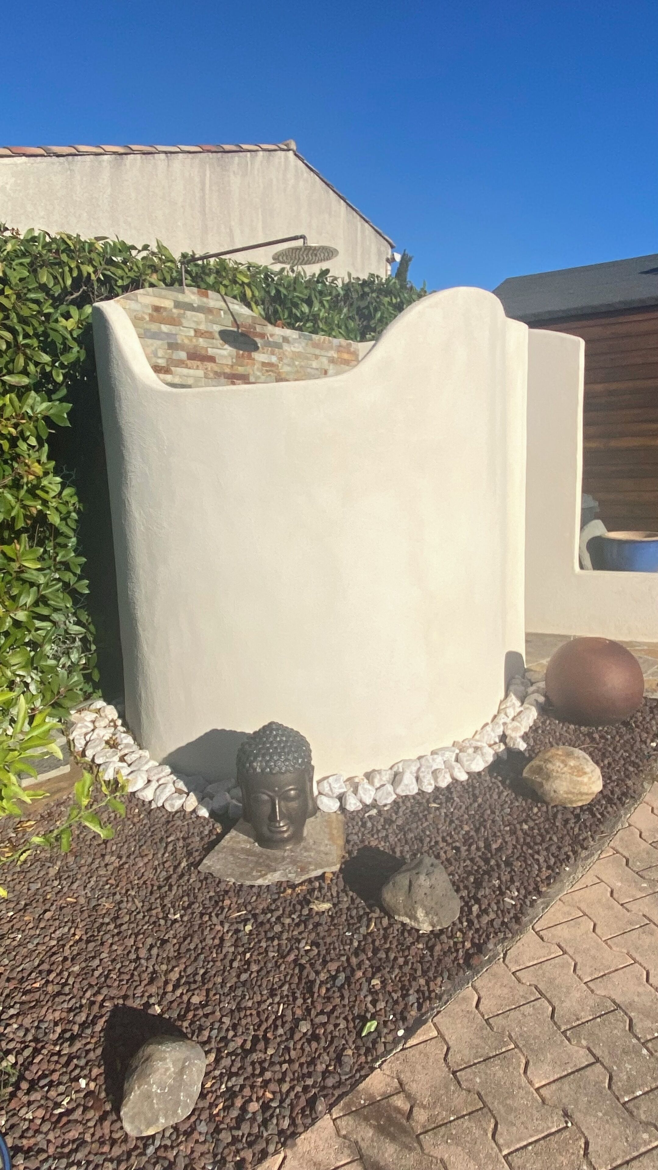 Un espace jardin agrémenté d’une statue de Bouddha, d’un sentier en pierre et d’un mur blanc orné d’une mosaïque.