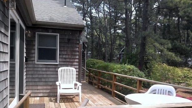 A wooden deck with a white chair and a table, surrounded by trees. - Pet Friendly Home, Short Walk to the Beach! (1807) (Wellfleet)