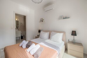 Quarto de hotel com cama, mesa de cabeceira, ar condicionado fixado na parede, estante com objetos decorativos e banheiro visível por uma porta aberta.