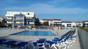 Une piscine entourée de chaises longues bleues et blanches, un bâtiment doté de balcons et un ciel dégagé.