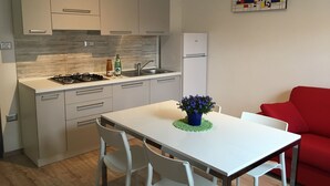 Ett modernt kök med vita skåp, en gasspis och en diskho. I matplatsen finns ett matbord med stolar och i vardagsrummet står en röd soffa.