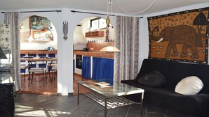 A living room with a black sofa, a glass coffee table, and a cowhide rug. There is a kitchen area with blue curtains and a mural of an elephant. - La casa del molino-Ideal for Two (Mogan)