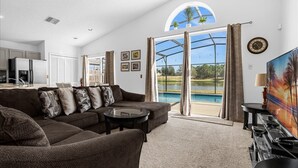 A modern living room with a sectional sofa, a glass coffee table, and a television. There is a large window with a view of a pool and greenery. - 4BR Kissimmee Home w/ Pool & Lake View – Near to Disney - A Family Favorite! (Kissimmee)
