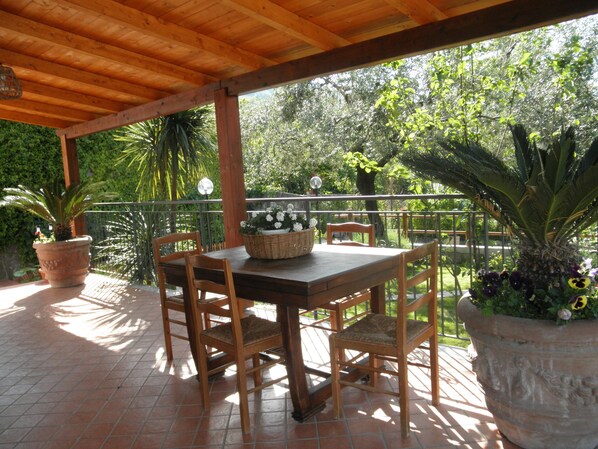 Uma mesa de madeira com cadeiras sob uma pérgola de madeira, cercada por plantas em vasos e um pátio com azulejos.