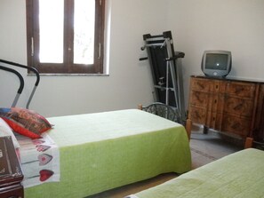 Quarto com duas camas de solteiro, cômoda de madeira, televisão por cima e janela com persianas.