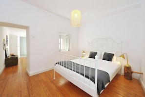 A bedroom with a white bed, a wooden floor, a hanging light fixture, and a mirror on the wall.