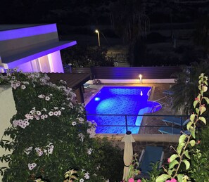 Una piscina con iluminación azul en su interior, rodeada de abundante vegetación y una terraza de madera.