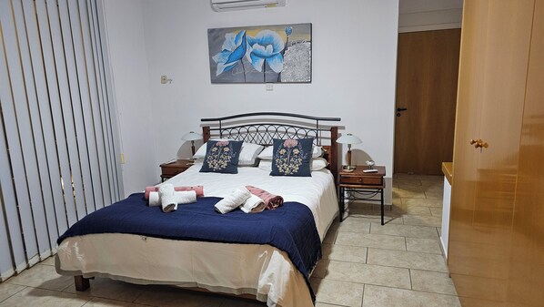 A neatly made bed with a blue blanket and pillows, a bedside table with a lamp, and a painting on the wall. - Luxury Villa With Private Pool and stunning sea views (Pissouri Bay)