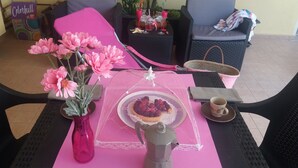 A table with a pink tablecloth, a vase of pink flowers, a lace-covered plate with a dessert, a coffee cup, and a teapot.