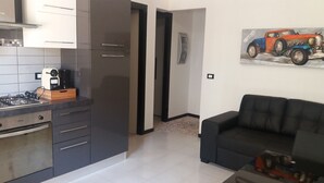 A modern kitchen with dark cabinets, a gas stove, and a coffee machine. There is a black sofa and a painting of a vintage car on the wall.