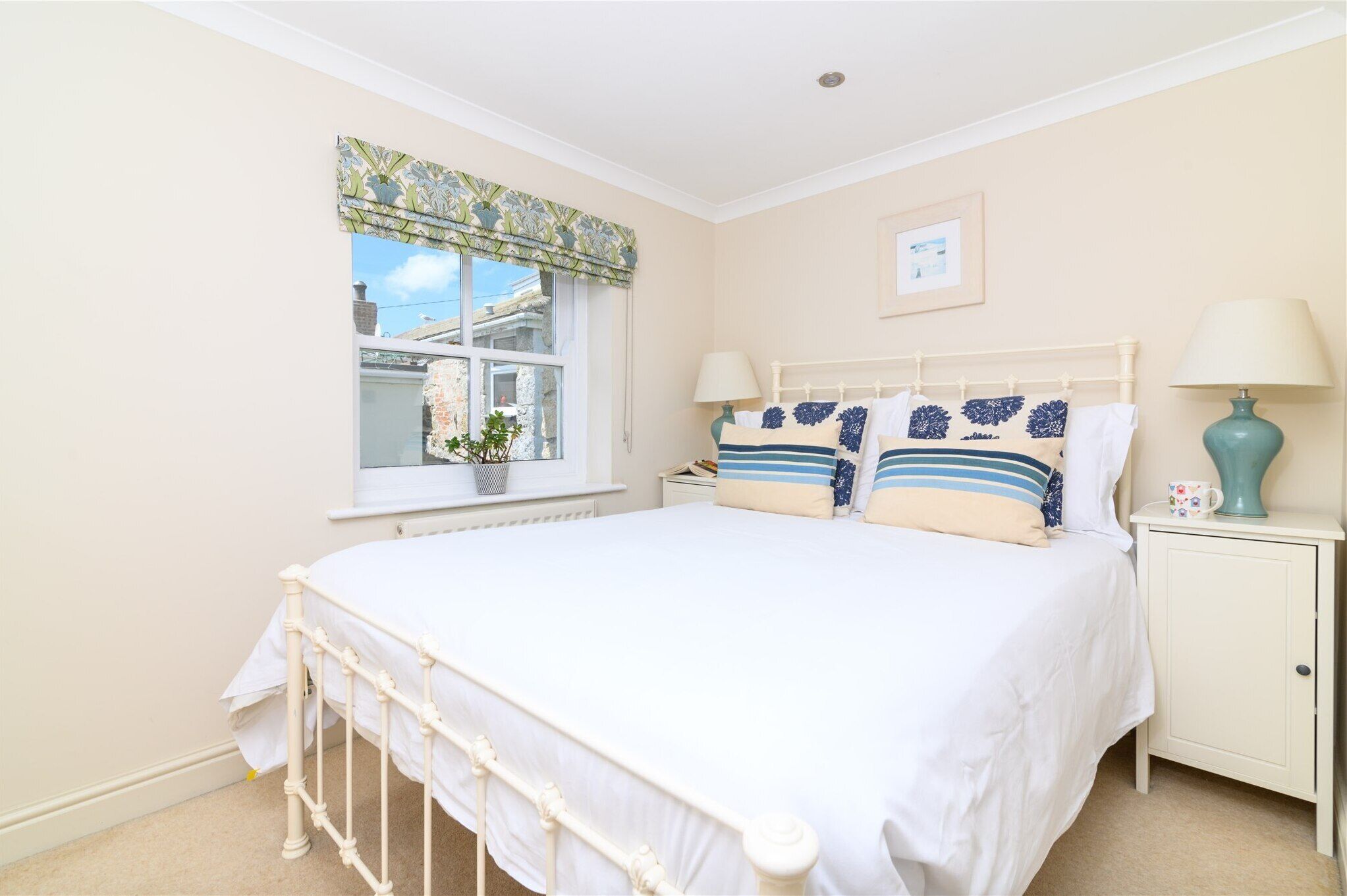 A bedroom with a bed, bedside lamps, a window with curtains, a small bedside table, and a picture on the wall.