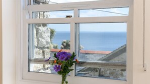 A window with a clear view of the sea and a vase with flowers on the windowsill. - Mousehole Cottage: sea views, parking & close to village centre, dog friendly. (Mousehole)