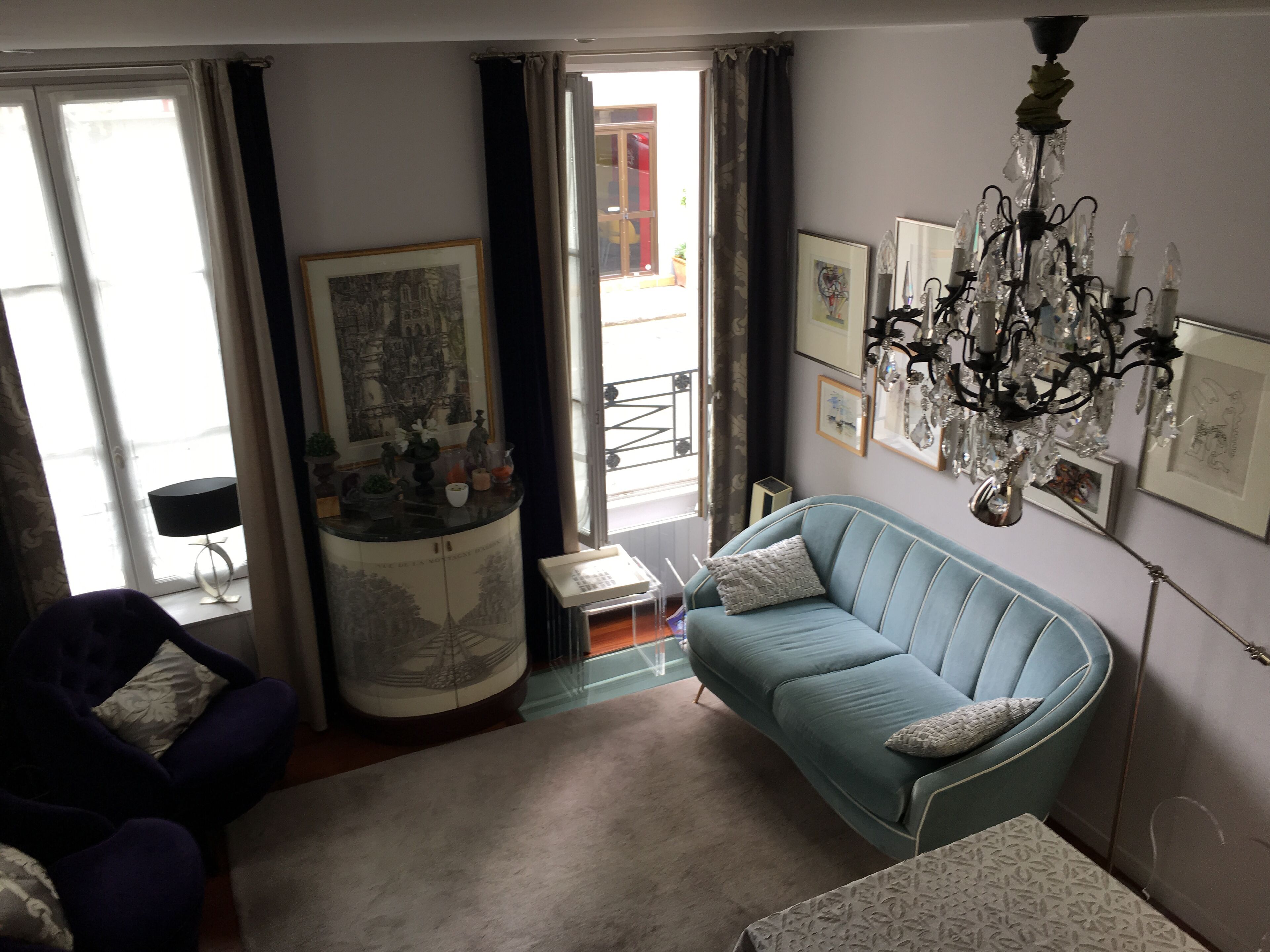 Een woonkamer met een blauwe bank, een witte fauteuil, een salontafel en een kroonluchter. Er hangen ingelijste foto’s aan de muur en een raam met gordijnen.