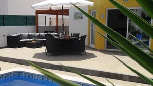 Uma piscina com borda azulejada, um guarda-sol, móveis de exterior e um edifício amarelo ao fundo.