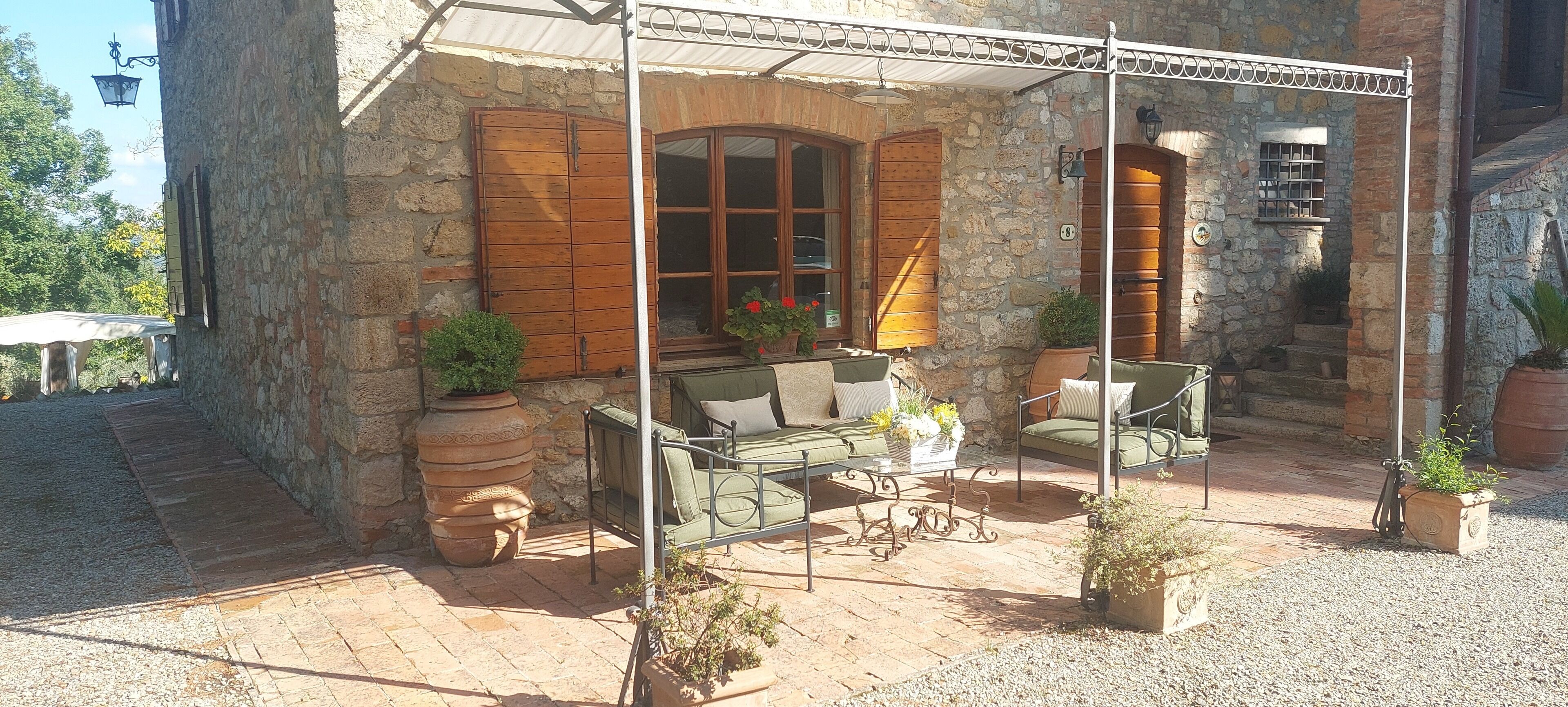 Ein Steinhaus mit überdachter Terrasse, möbliert mit einer Couch und Sesseln, und mit Blumentöpfen.