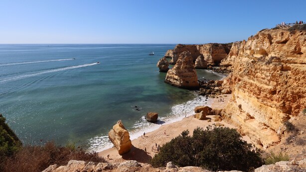Un paysage côtier caractérisé par une falaise rocheuse, une mer d’un bleu limpide et une plage de sable.