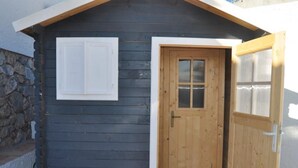 A small wooden cabin with a dark exterior, a white door with glass panels, and a stone wall to the left.
