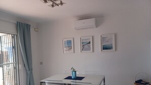 Quarto de hotel moderno com uma mesa branca, cadeiras transparentes, um vaso azul e três quadros na parede.