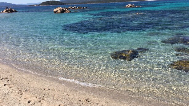 Une plage bordée d’une eau turquoise et limpide, de formations rocheuses et dominée par des montagnes lointaines.