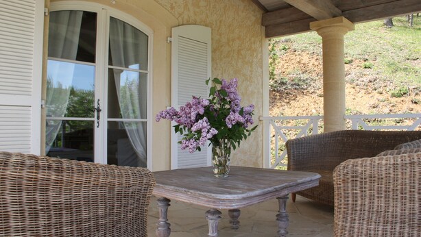En overdækket terrasse med fletningsmøbler, et bord med en blomstervase og udsigt til en skråning.