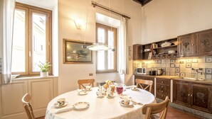 Un comedor con una mesa redonda dispuesta para cuatro personas, rodeada de sillas de madera. Un mantel blanco cubre la mesa, y hay platos, tazas y una jarra de jugo encima. La habitación tiene armarios de madera, una lámpara colgante y una ventana con cortinas.