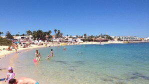 Ein Strand mit klarem Wasser, Menschen beim Schwimmen und Sonnenbaden, und Gebäuden im Hintergrund.