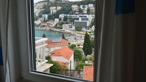 A coastal cityscape with buildings, a beach, and a view through a window with curtains. - Studio apartment Julia with balcony in Lapad (Dubrovnik)