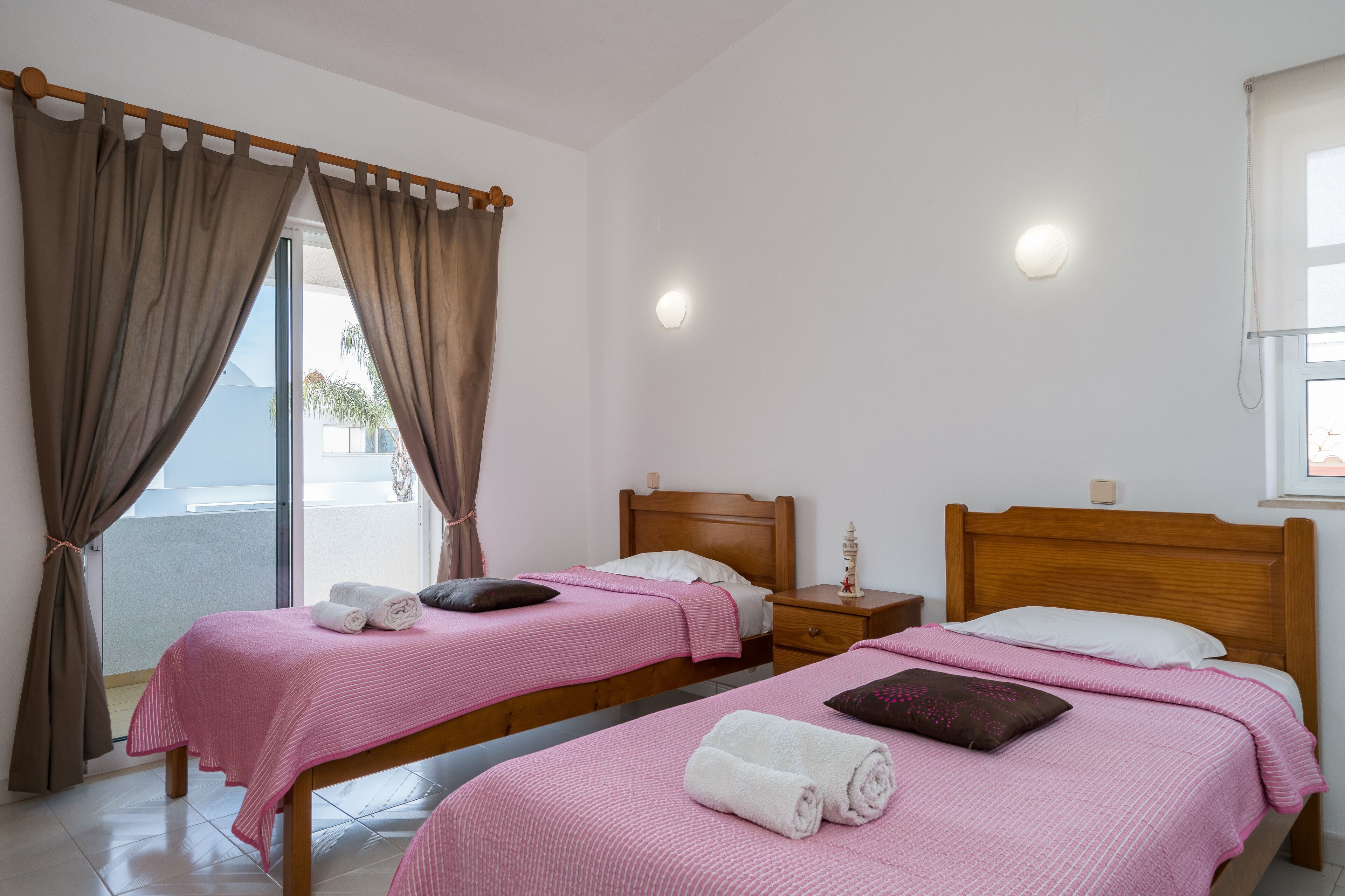 Een hotelkamer met twee bedden, elk voorzien van roze beddengoed en kussens, een houten hoofdbord, een nachtkastje en een raam met gordijnen.
