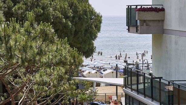 Ein Balkon mit Meerblick, ein Strand mit Sonnenschirmen und ein Baum.