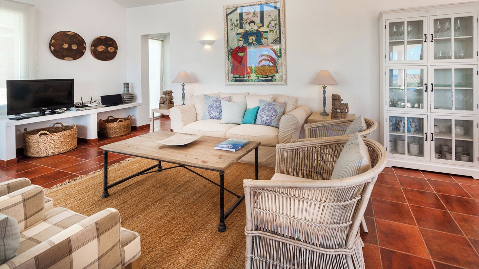 A living room with a sofa, coffee table, wicker chairs, a television, and a cabinet with glass doors.