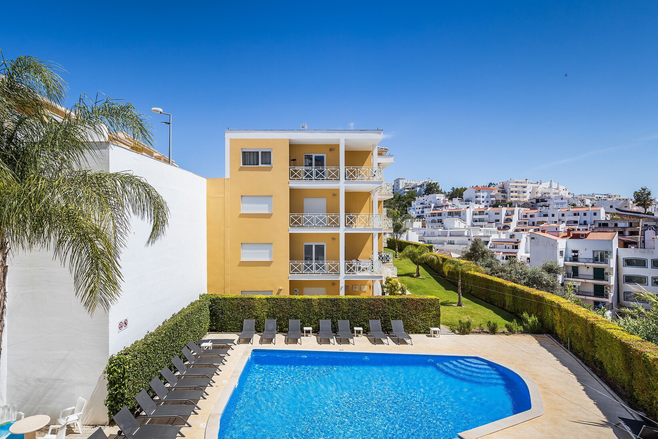 Un edificio amarillo con piscina, sillas de descanso y vistas a una zona residencial en una colina.