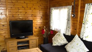 A wooden cabin interior with a television on a wooden stand, a sofa with patterned cushions, and a vase with flowers. - Log Cabins In The Beautiful Highlands Of Zeliv, By A Lake and forest. Oak cabin (Zeliv)