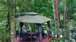 A wooden deck with outdoor furniture, surrounded by trees and overlooking a body of water. - Log Cabins In The Beautiful Highlands Of Zeliv, By A Lake and forest. Oak cabin (Zeliv)