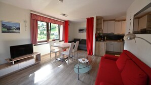 Una sala de estar moderna con un sofá rojo, una mesa de centro de vidrio, un televisor sobre un mueble de madera, un área de comedor con mesa y sillas, y una cocina con armarios y una encimera.