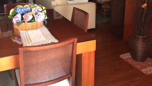 Un coin repas avec une table en bois, des chaises et un arrangement floral en guise de décoration centrale. On aperçoit un canapé et une bibliothèque à l’arrière-plan.