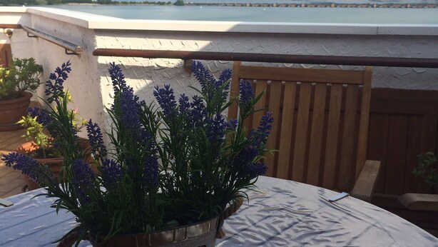 Une table sur laquelle se trouve un seau en métal étiqueté « jardin », contenant des plants de lavande, est placée sur un balcon donnant sur un lac et des montagnes.