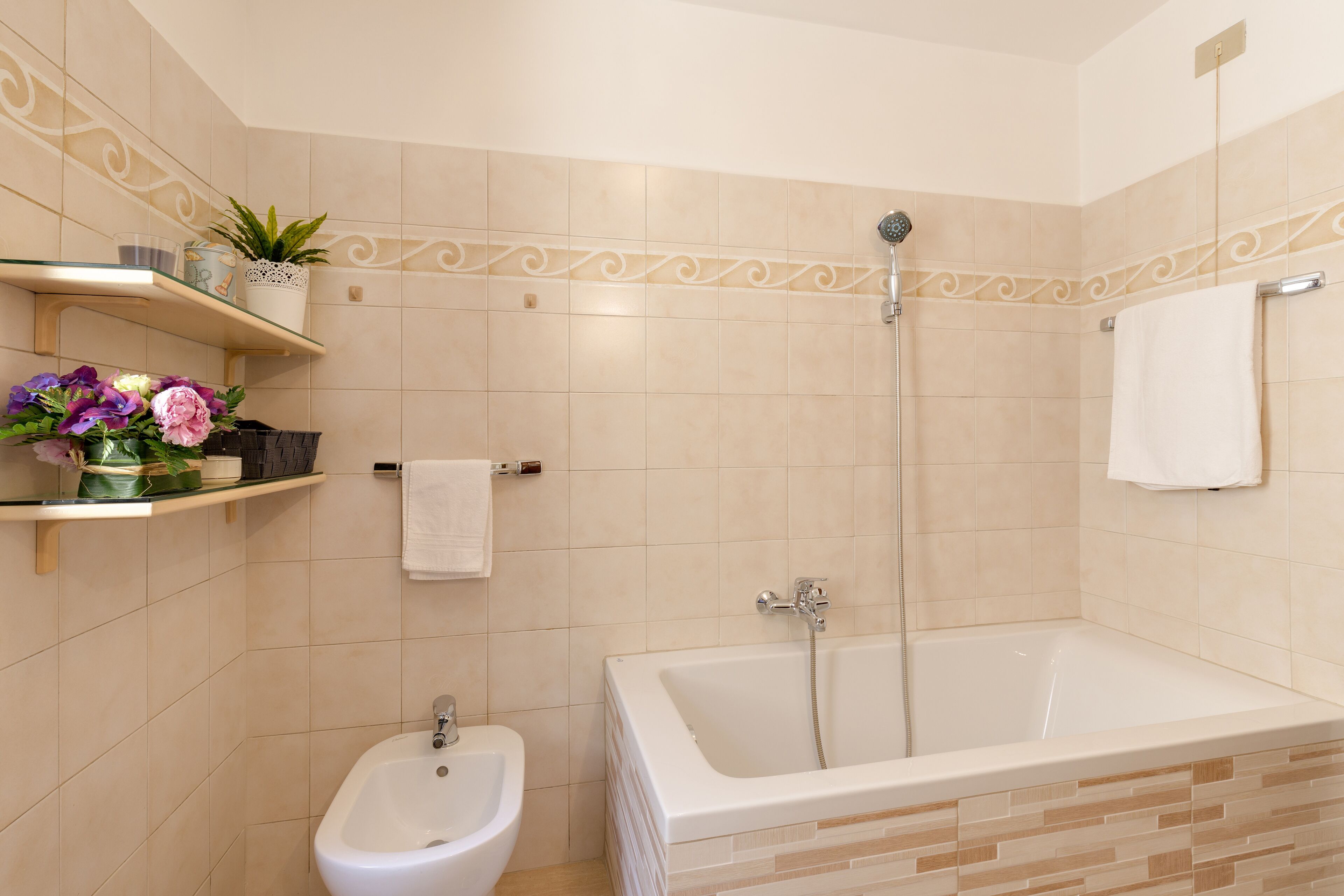 Een badkamer met een bad, een hygiëneschuimer en een douche. Er zijn planken met toiletartikelen en een potplant.