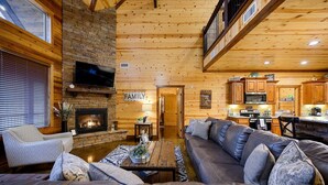 A cozy log cabin interior with a fireplace, a large leather sofa, a television, and a kitchen area. - Luxury Broken Bow Cabin w/ Game Room & Hot Tub | Sleeps 16 (HOCHATOWN)